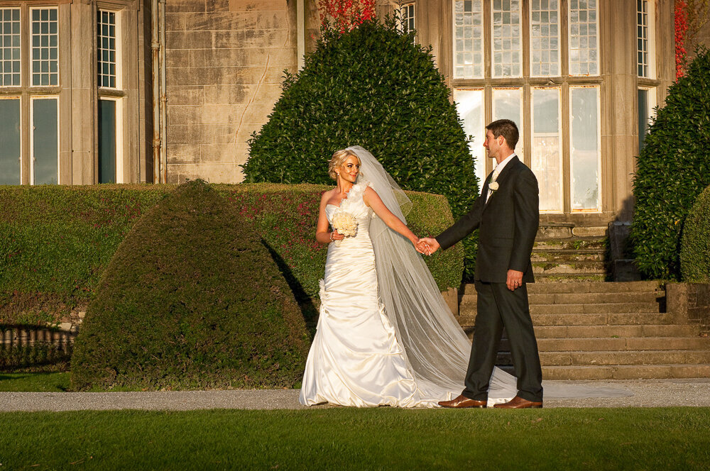 Bride in satin mermaid wedding dress and long veil, holding white flower bouquet, holding hands with groom wearing a black wedding suit at Muckross House
