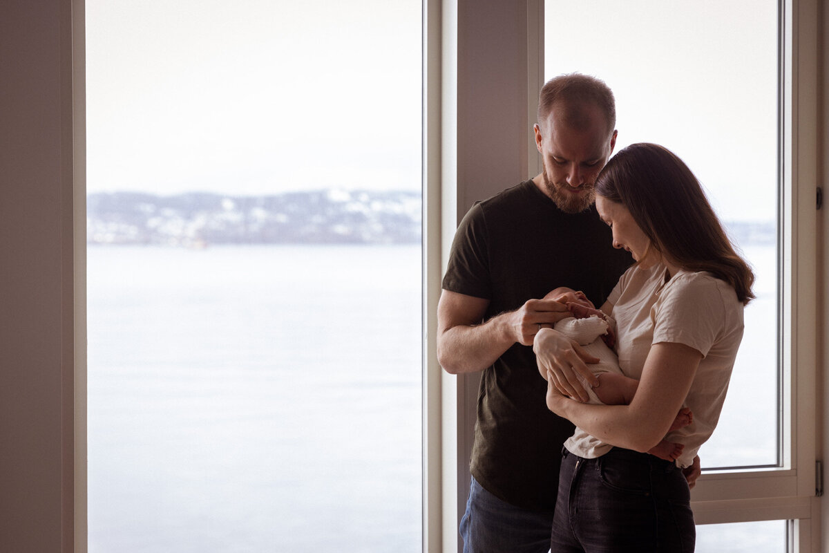 Livsstilsfoto familieportrett tatt ved vindu. Far og mor står ved vindu, mor holder baby, far stryker baby på hodet.