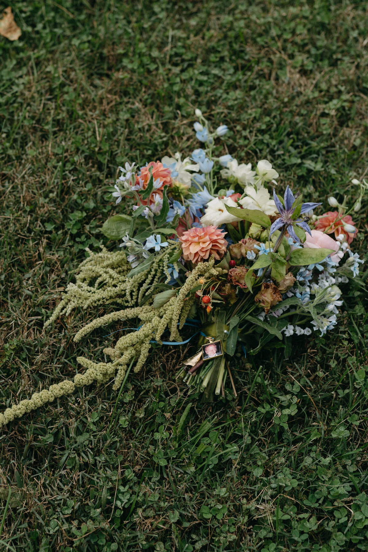 Lush garden-style bridal bouquet resting on grass, featuring pastel dahlias, roses, delphinium, greenery, trailing amaranthus, and a sentimental photo charm tied to the bouquet stems.
