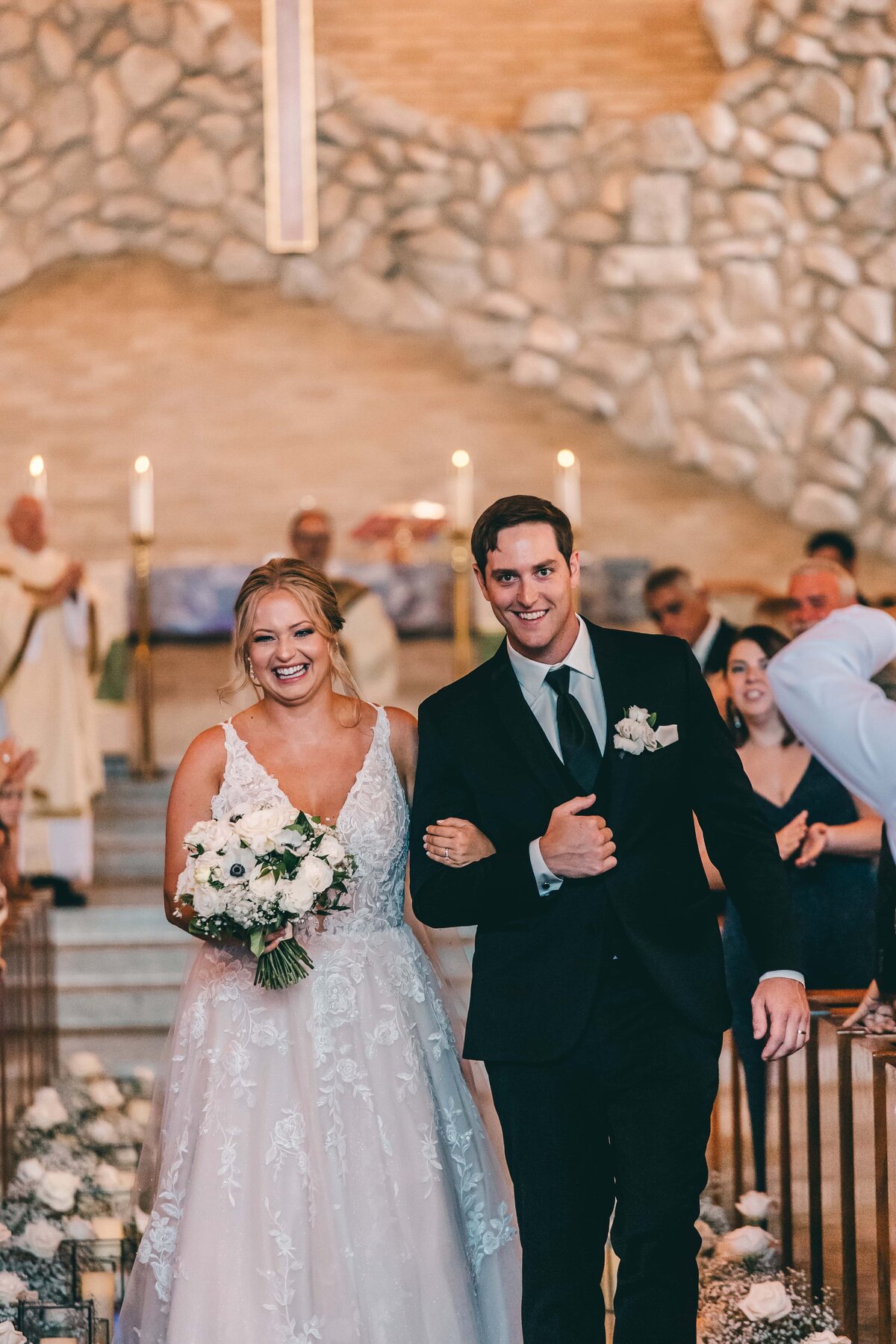 A joyful bride and groom walking down the aisle at a park farm winery wedding, smiling broadly, with the bride holding a bouquet and the groom in a black suit.