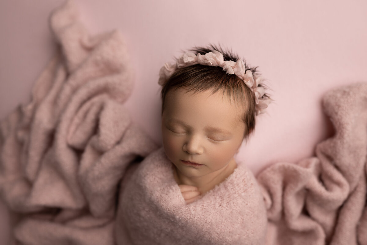 Philadelphia's best newborn photographer, Katie Marshall captures a sleeping baby. The baby is swaddled in dusty rose with her fingers peeking out from the swaddle. She is wearing a matching headband adorned with ruched bows.