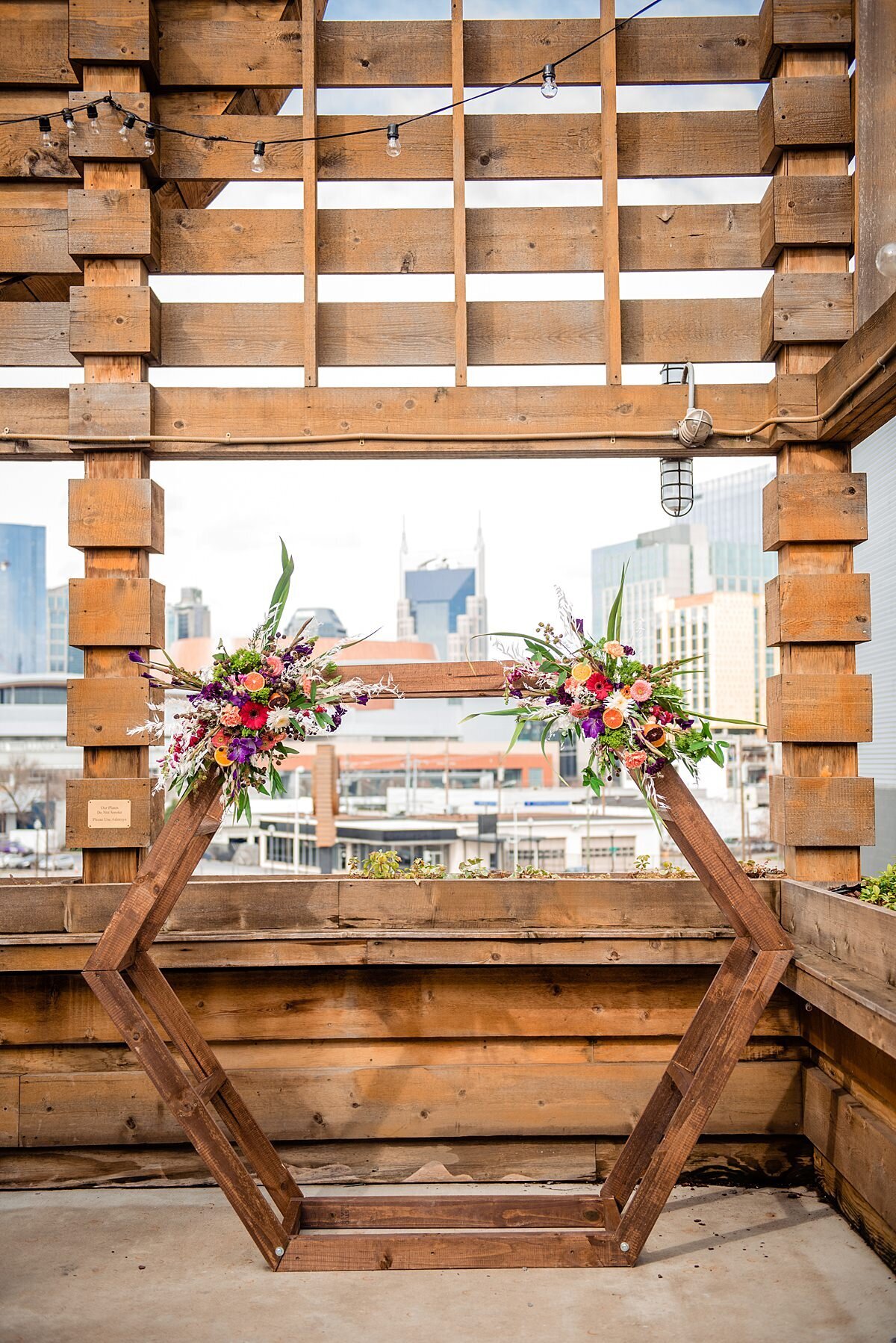 Octagonal barn wood wedding ceremony arbor  sits on the patio at City Winery Nashville. The Nashville city skyline is in the background. The circular arbor is decorated with two floral sprays of red, white, pink and purple flowers with eucalyptus.