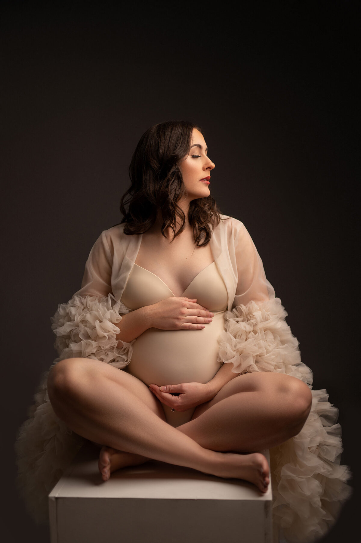 Maternity portrait of a barefoot woman seated cross-legged on a pedestal. She is wearing a nude body suit and matching sheer, ruffled robe with her eyes closed and head turned to her left.