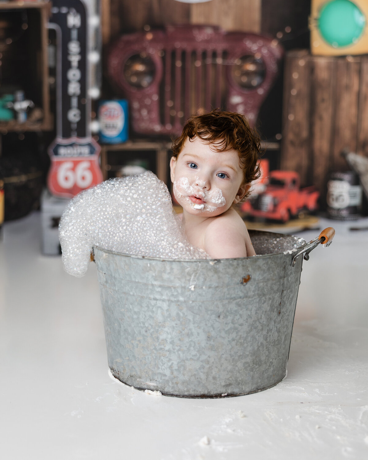 Baby boy with curly hair smiling in a galvanized tub filled with bubbles, foam on his face, set against a vintage garage-themed backdrop with Route 66 signs. Photograph by Yucaipa cake smash photographer Kaitlyn Dawn Photography.