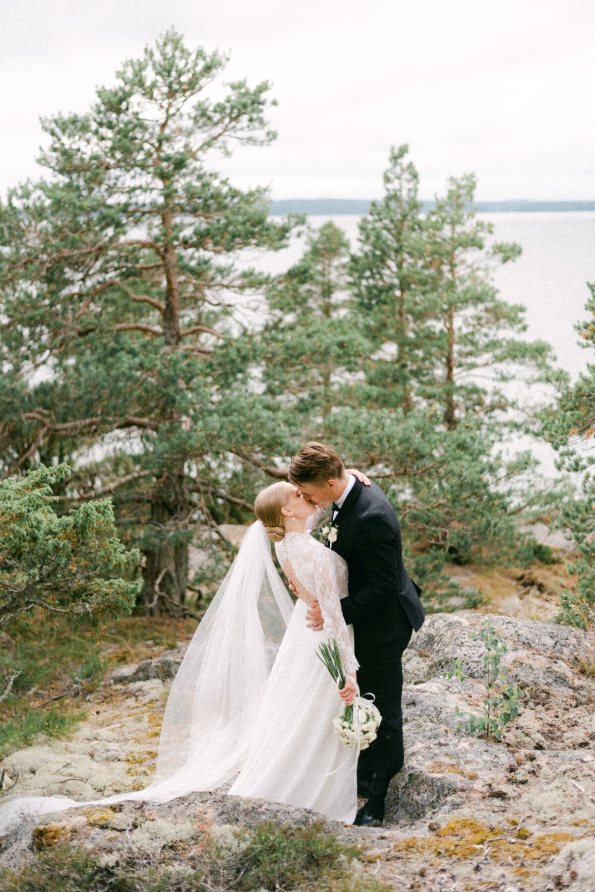 A wedding couple is kissing in the forest by the sea in a photograph taken by wedding photographer Hannika Gabrielsson in Finland.