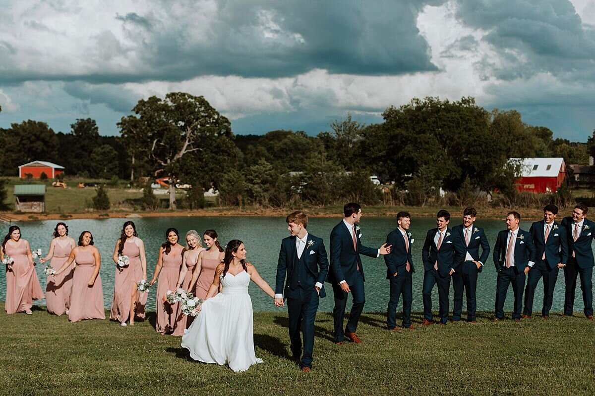 bridesmaids in blush dresses and groomsmen in black suits with bride and groom lakeside