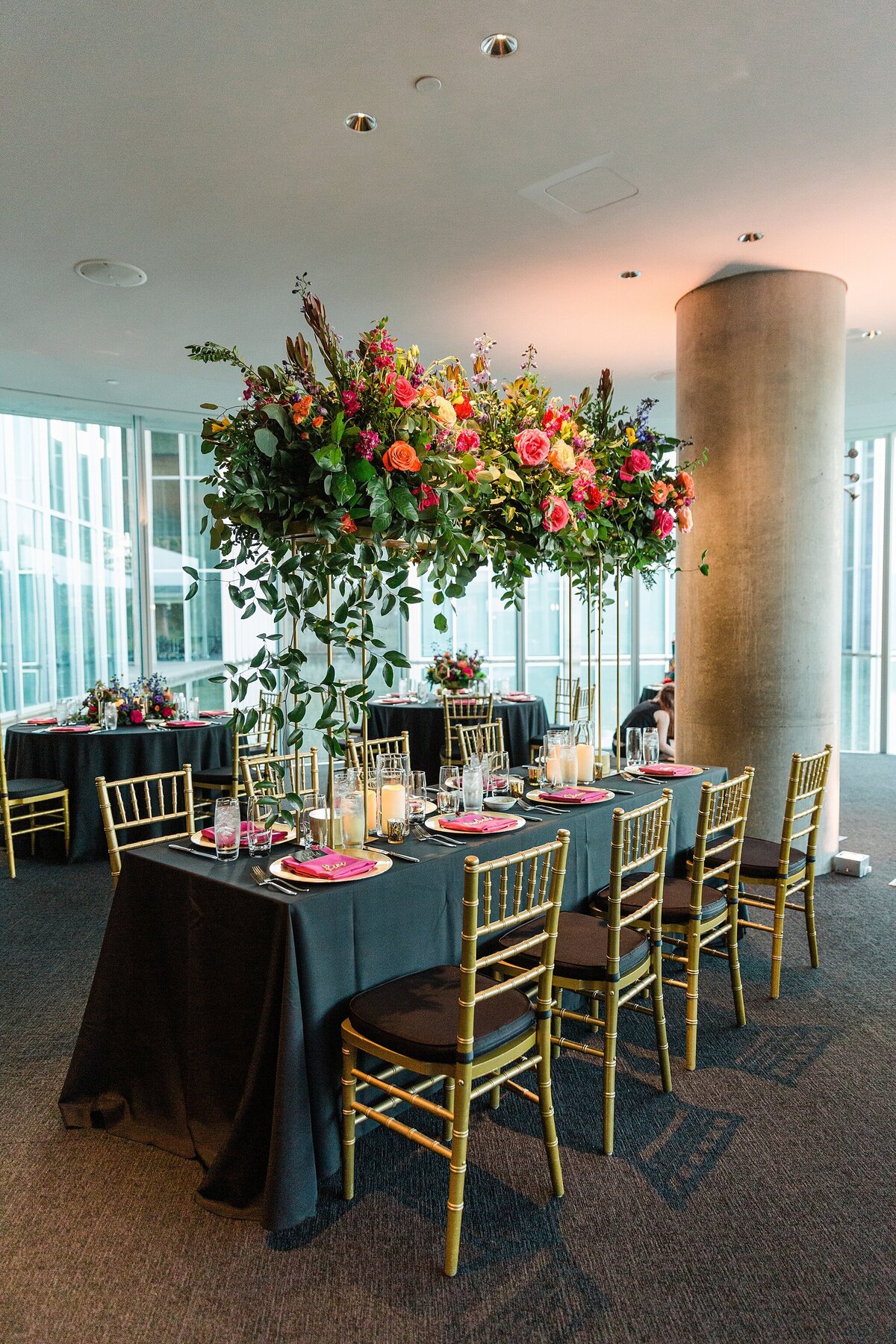 A detail shot of a reception table at a wedding at the Modern Art Museum of Fort Worth in Fort Worth, Texas. The rectangular table is draped in a long black tablecloth and covered with place settings, candles, and a large, tall floral centerpiece. The table is surrounded by eight gold and black chairs.