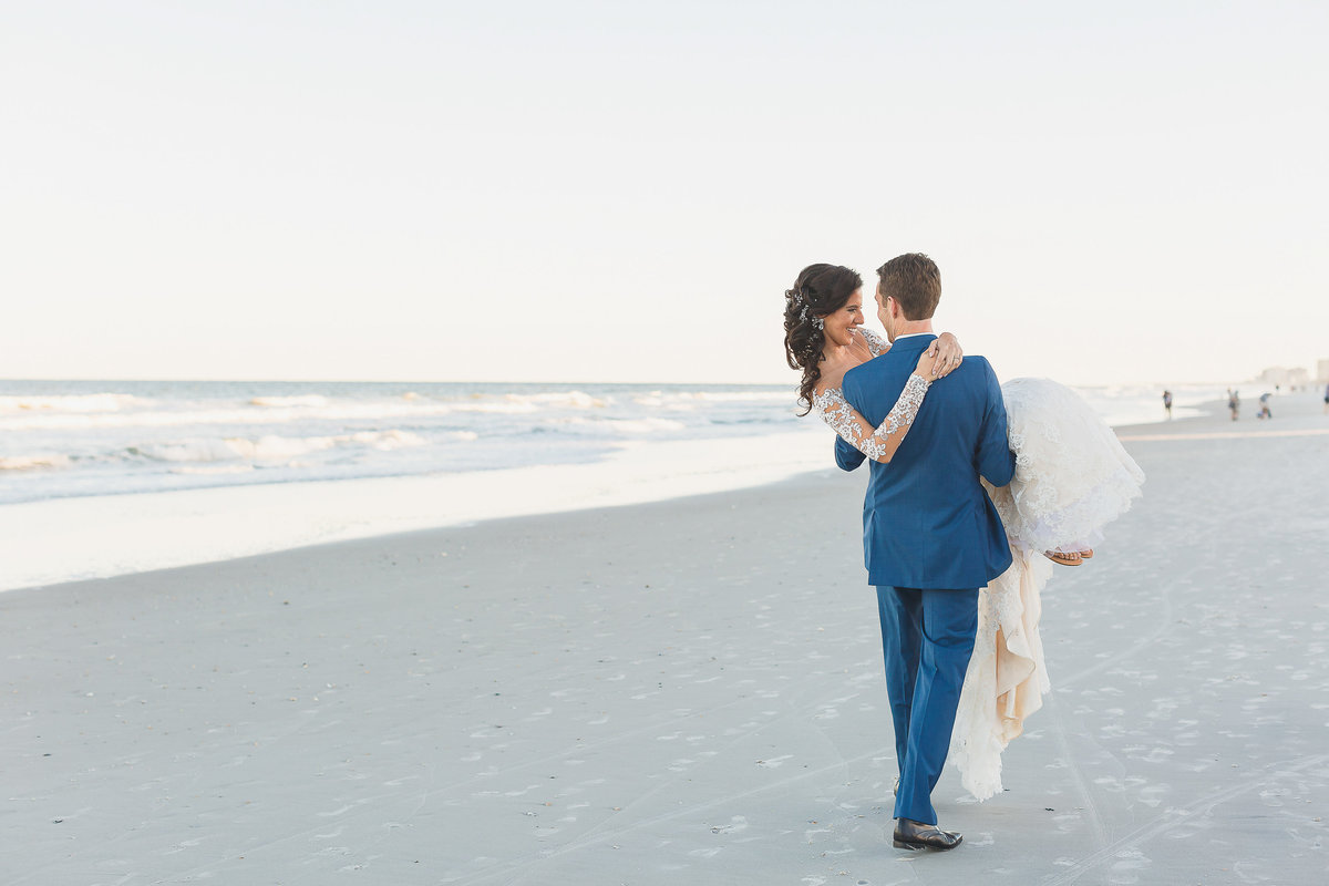 Groom in blue suit carries bride across the sand after their Atlantic Beach wedding.