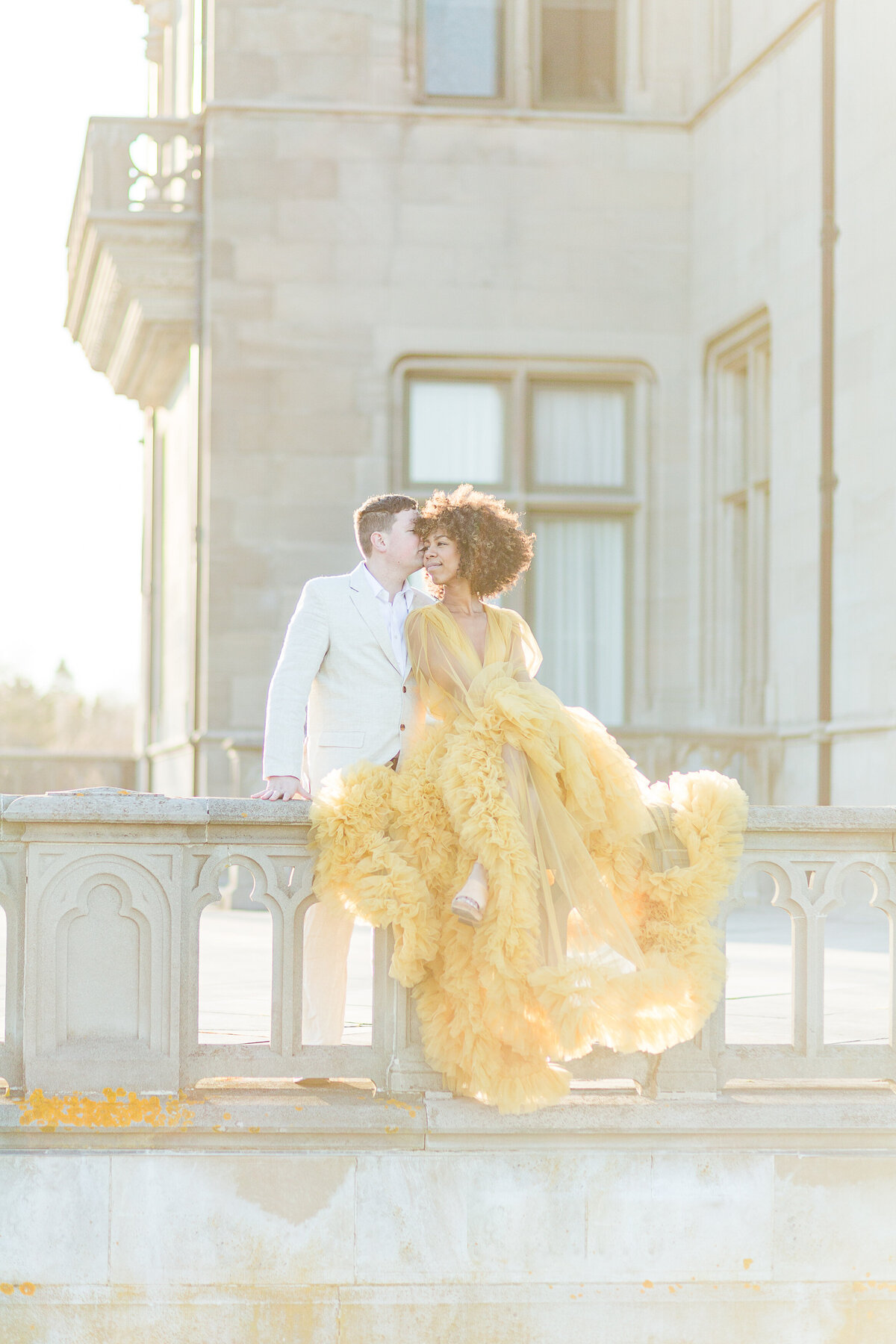 Man and woman are sitting on Salve Regina's stone walls for their sunset engagement photoshoot. The woman is in an elaborate yellow organza gown and the man in a white suit. The woman is looking toward the man who is gently kissing her on the cheek. Salve Regina's buildings are featured prominently in the background. Captured at golden hour creating a soft haze. Photo by Lia Rose Weddings, best Rhode Island Wedding and Engagement Photographer
