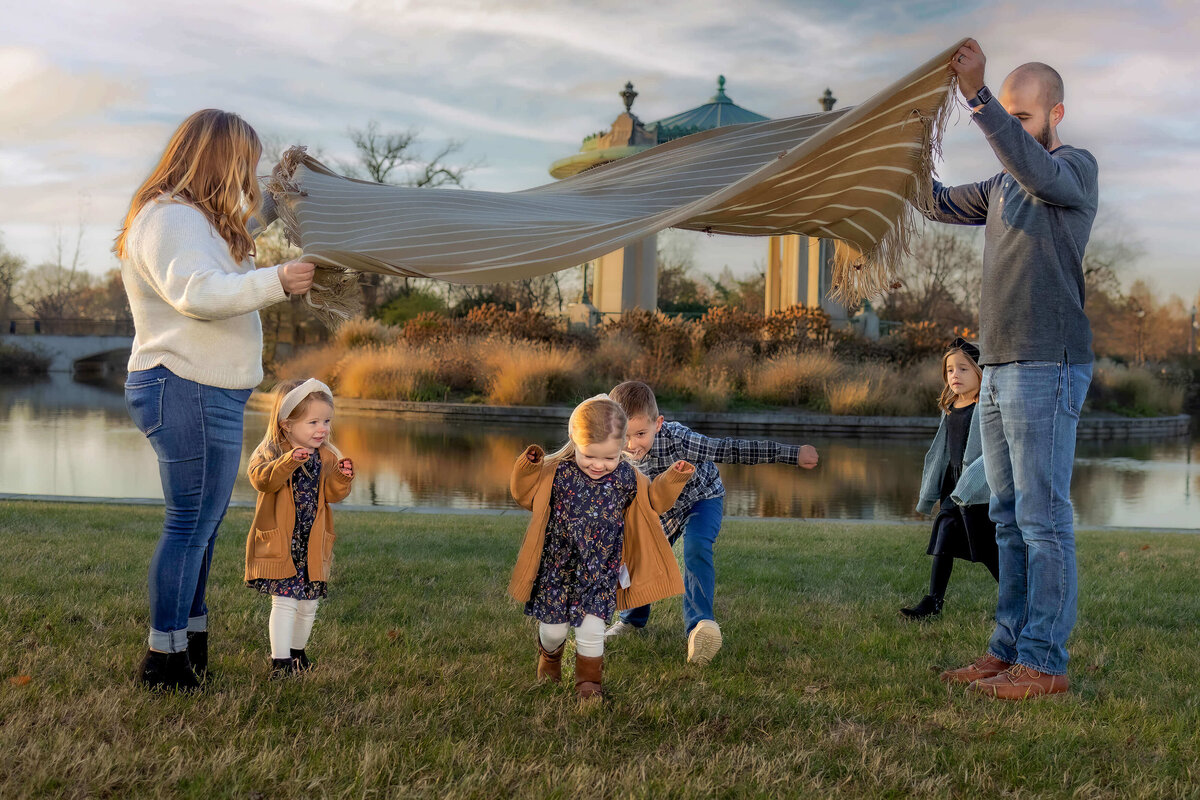 4 young kids running under a blanket while mom and dad fluff it into the air by a pond at sunset