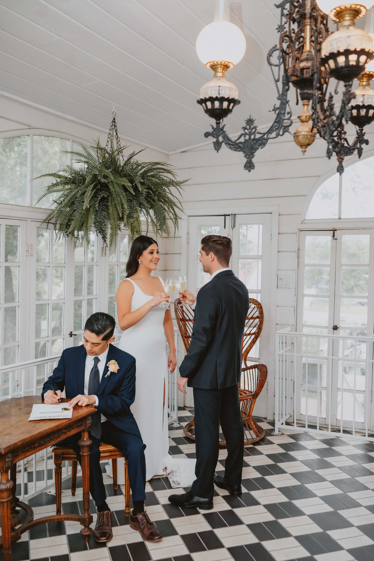 Bride and groom share a champagne toast indoors while the officiant signs the marriage license at a vintage venue with black-and-white tile floors and elegant chandeliers.