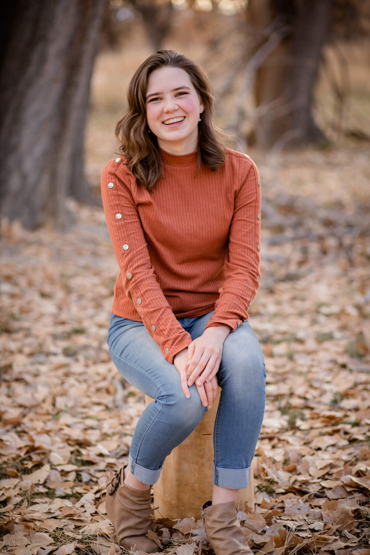 teenage girl wearing a rust colored sweater and jeans sitting on a log surrounded by fallen leaves