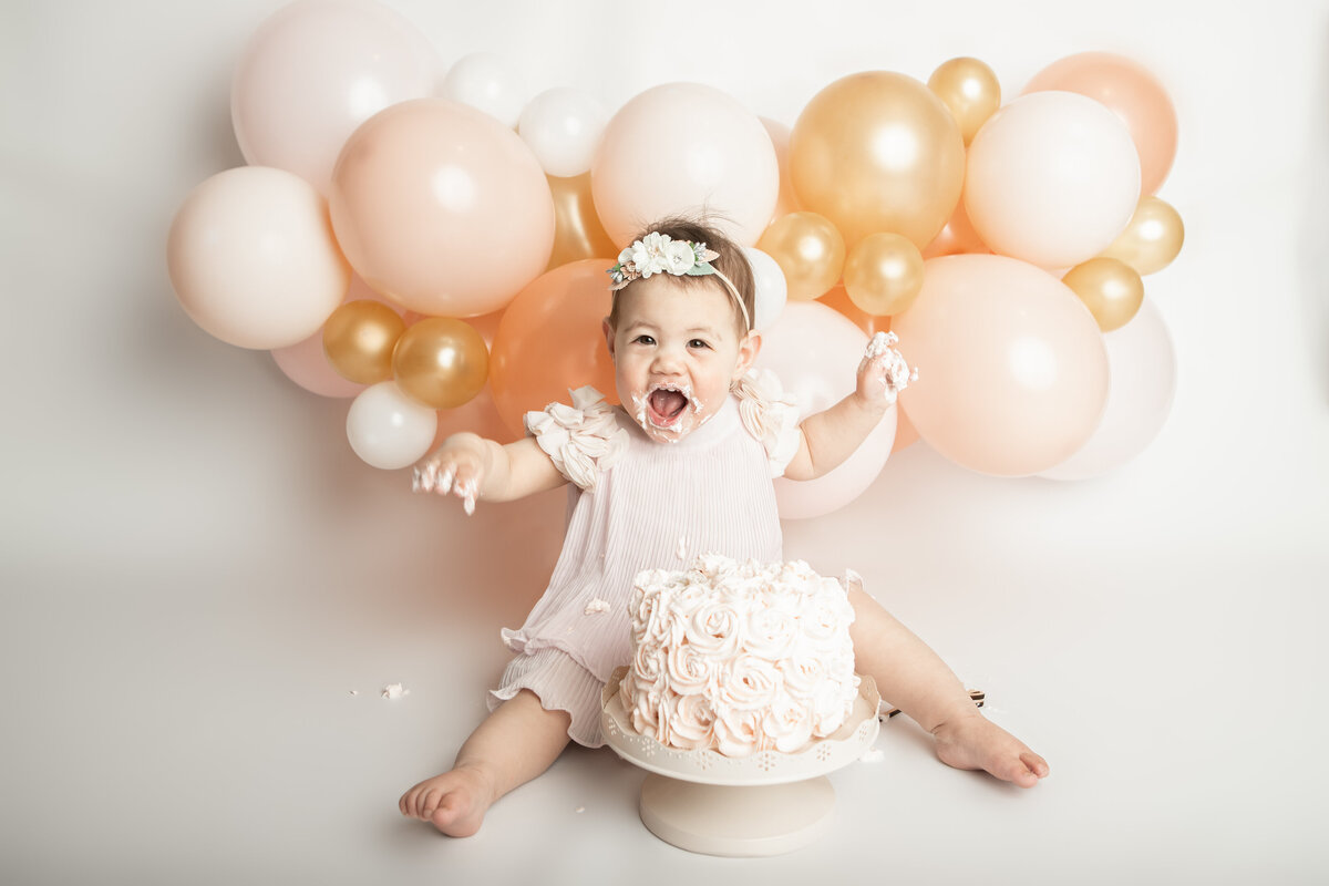 Birthday baby girl eating a messy pink cake with a pink peach white and gold balloon garland