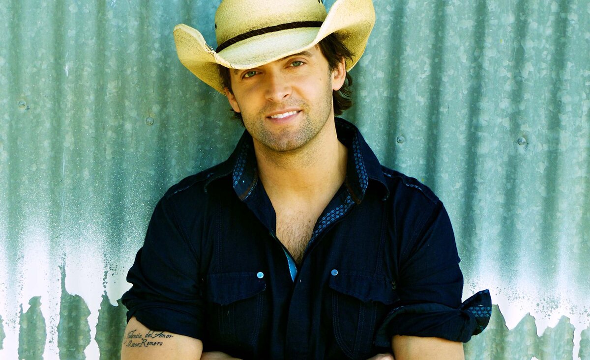 Country music photo Dean Brody wearing straw cowboy hat blue shirt leaning against green metal wall