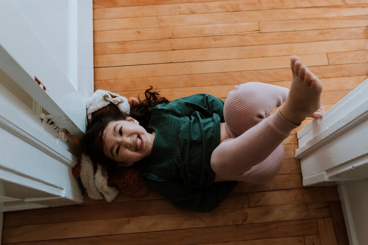 Family Photographer, a young girl lays on the hardwood floor smiling