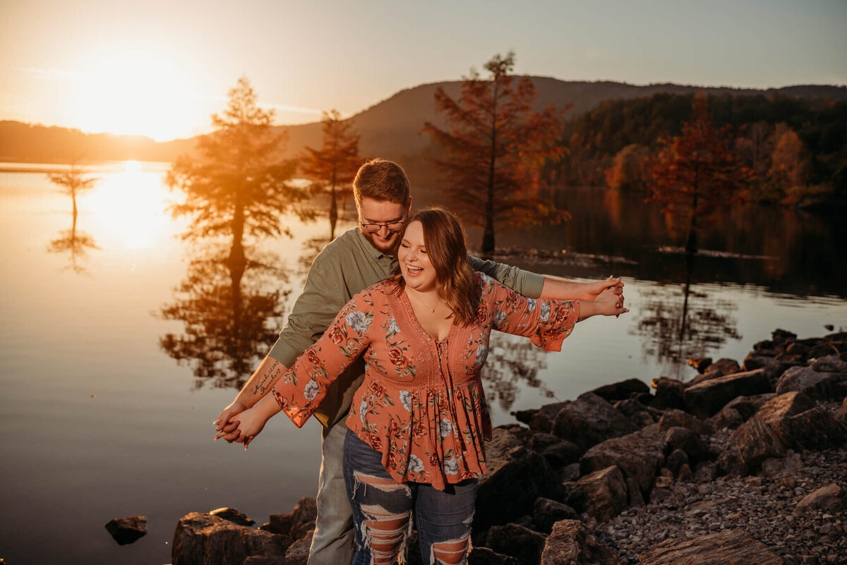 photo of a woman and man laughing in front of a lake with trees in it at sunset