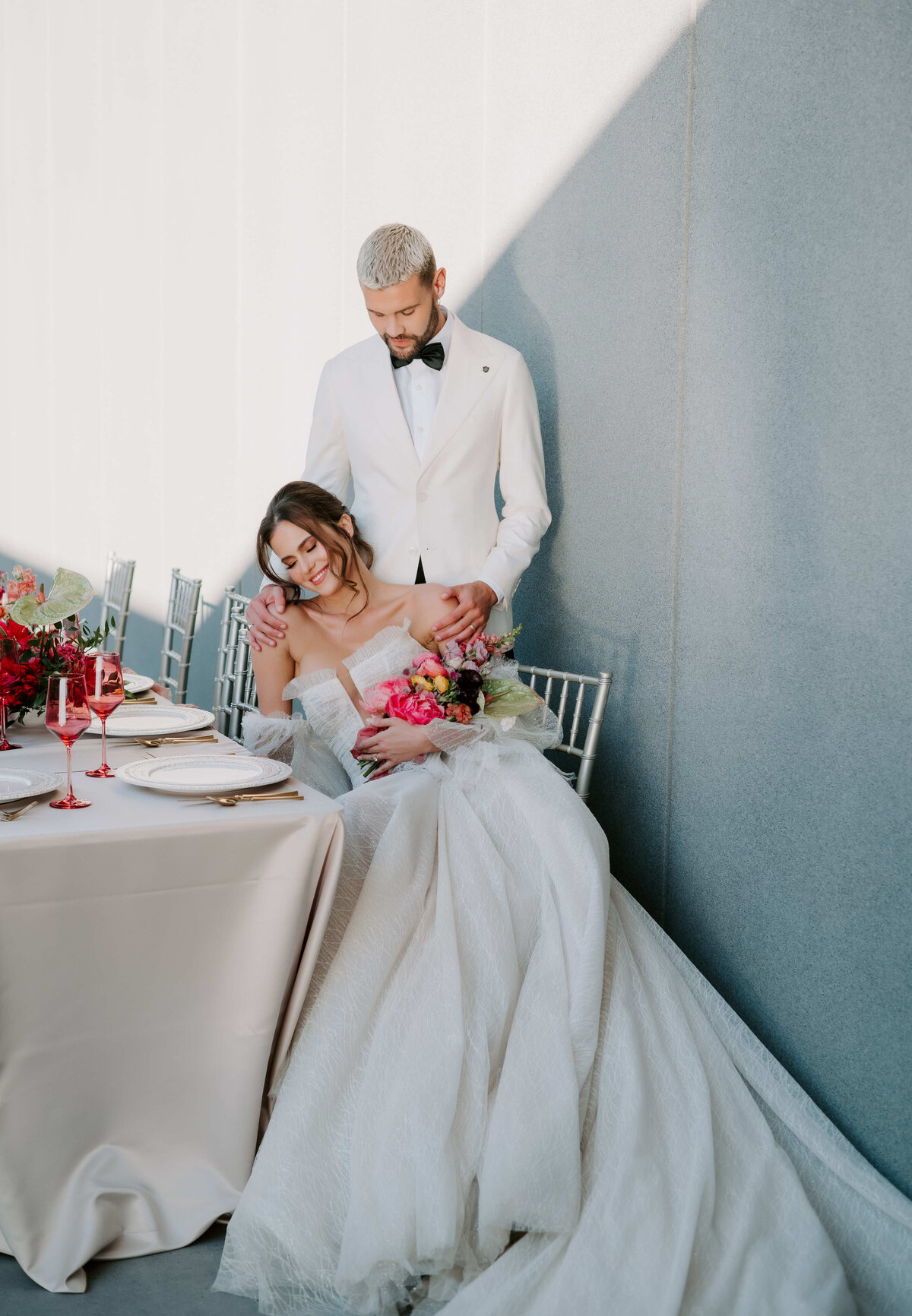 Bride in elegant white gown sits at a table adorned with red flowers, holding a bouquet. Groom in white suit stands behind, creating a romantic scene.