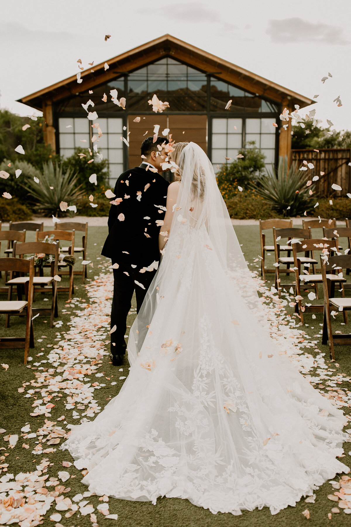 A bride and groom standing in the middle of an aisle kissing with flower petals surrounding them