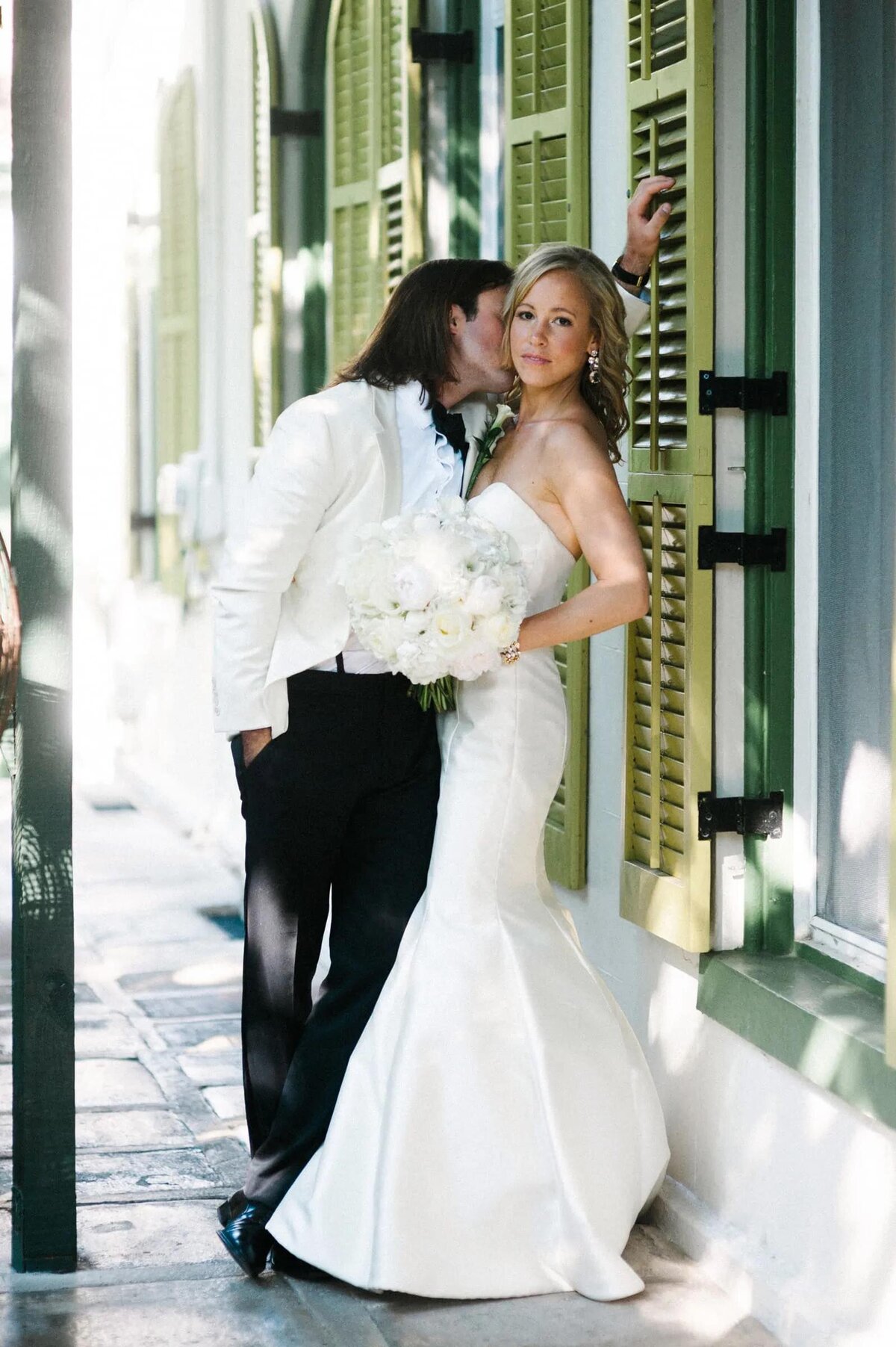 A bride leans against a wall in an elegant, form-fitting wedding gown, receiving a kiss from her groom on a sunlit street
