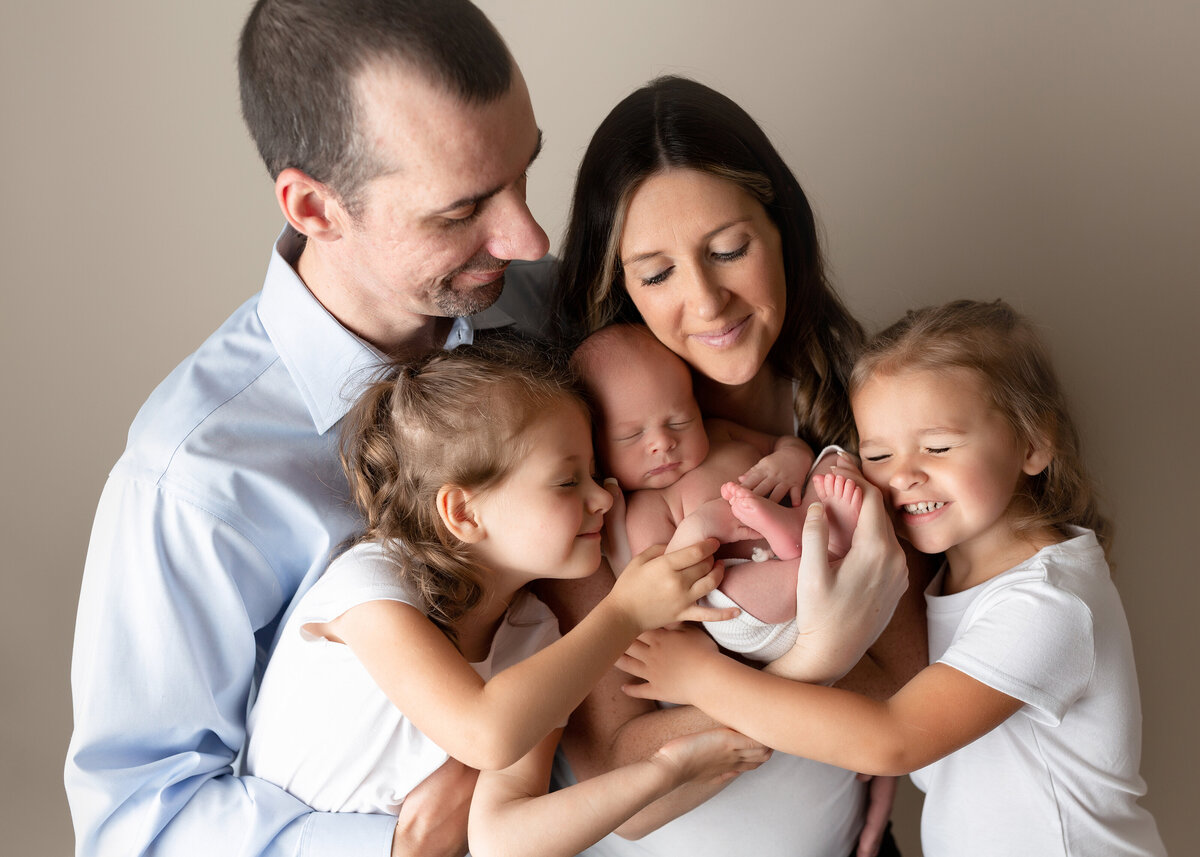 Family newborn photos in West Palm Beach and Jupiter newborn photography studio. Mom, dad, and two big sisters are cuddling baby with their eyes closed. Baby is in the center of the image with his family surrounding him.