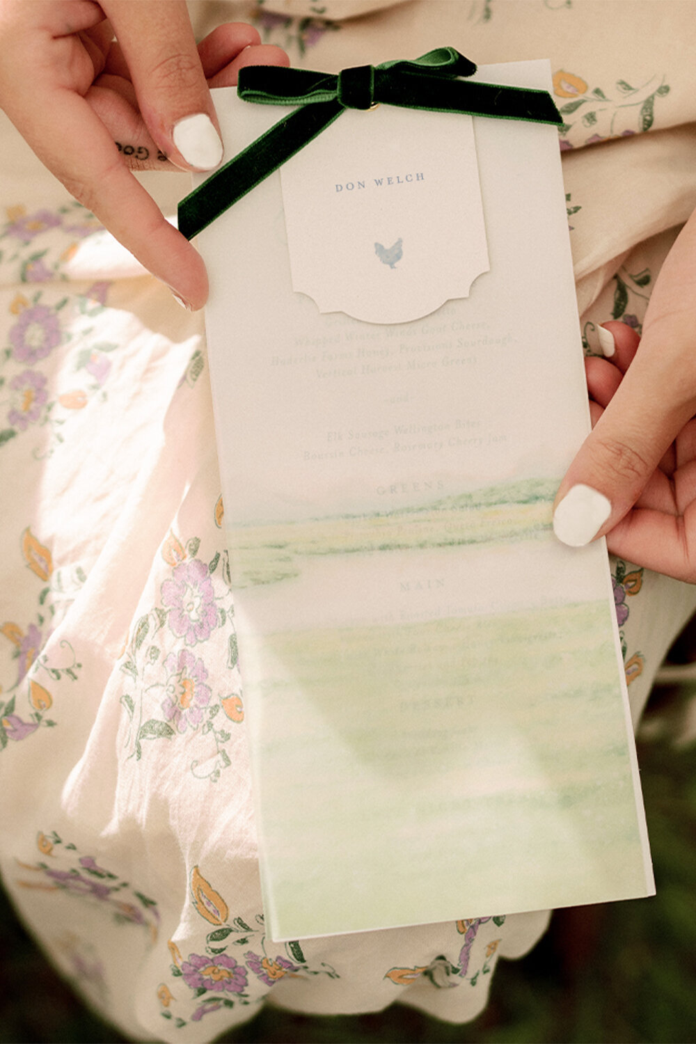 Wedding guest holding an elegant menu with a translucent vellum overlay, featuring an illustration of the venue. The menu is tied with a luxurious green velvet ribbon.