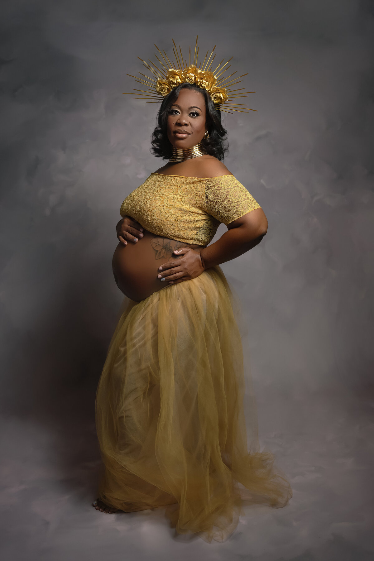 A mother to be in a gold gown and gold headpiece holds her bump in a studio