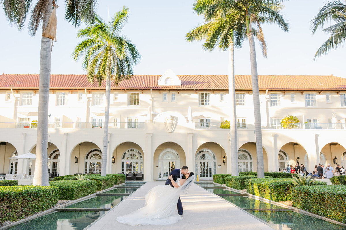 A couple shares a kiss at the center of a narrow water feature in front of an elegant building with arched doorways and palm trees, captured by their destination wedding photographer.