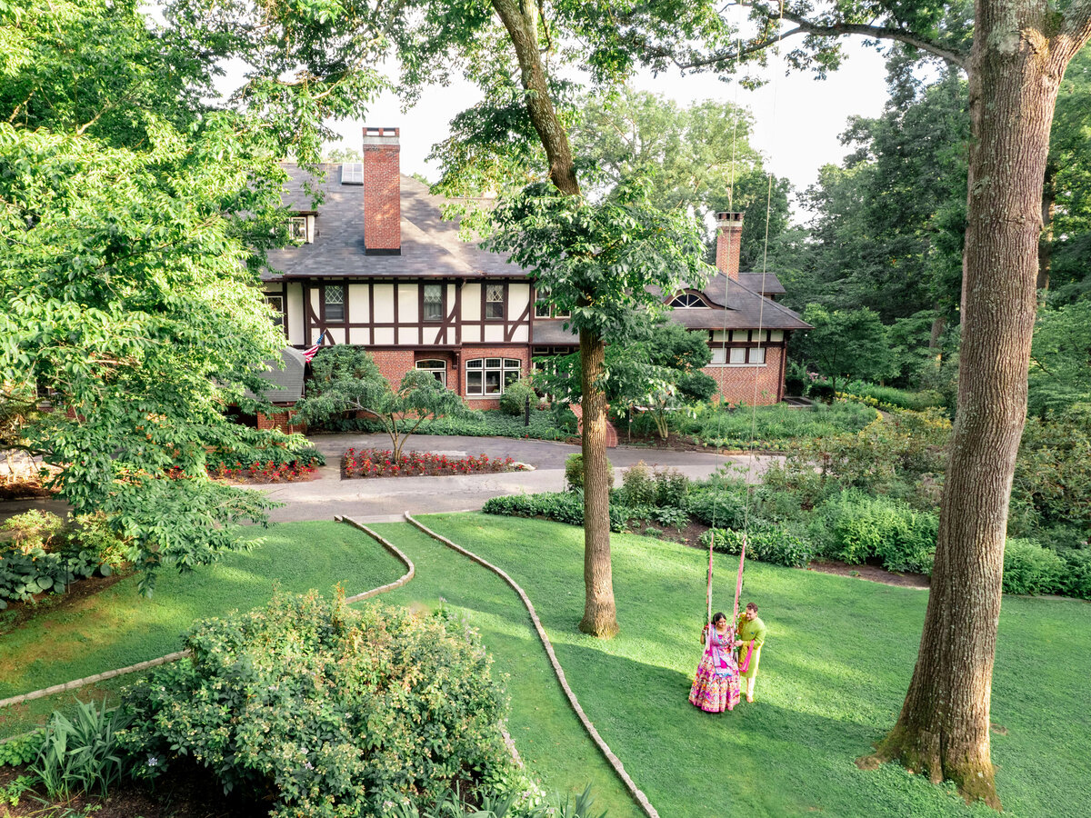 A large, two-story Tudor-style house surrounded by lush greenery and gardens. Two children play on a swing hanging from a tree in the foreground. The scene is serene, capturing a sunny day with expansive lawns and mature trees.