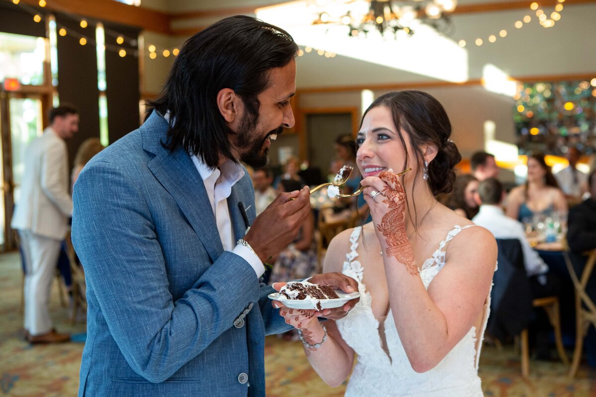 A bride and groom feeding each other cake at their Iowa wedding reception, surrounded by guests. The bride has henna patterns on her hands.