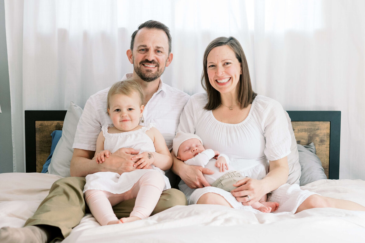 A new family of 4 sit on a white bed in a white room and smile at the camera of Virginia family photographer