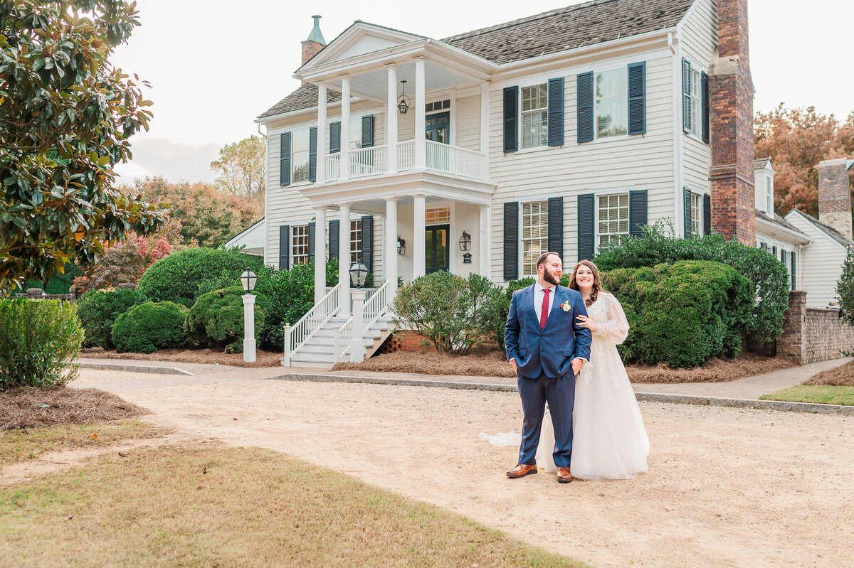 A smiling bride and a groom in a blue tux standing outside of The Sutherland on their fall wedding day by JoLynn Photography, a Raleigh wedding photographer