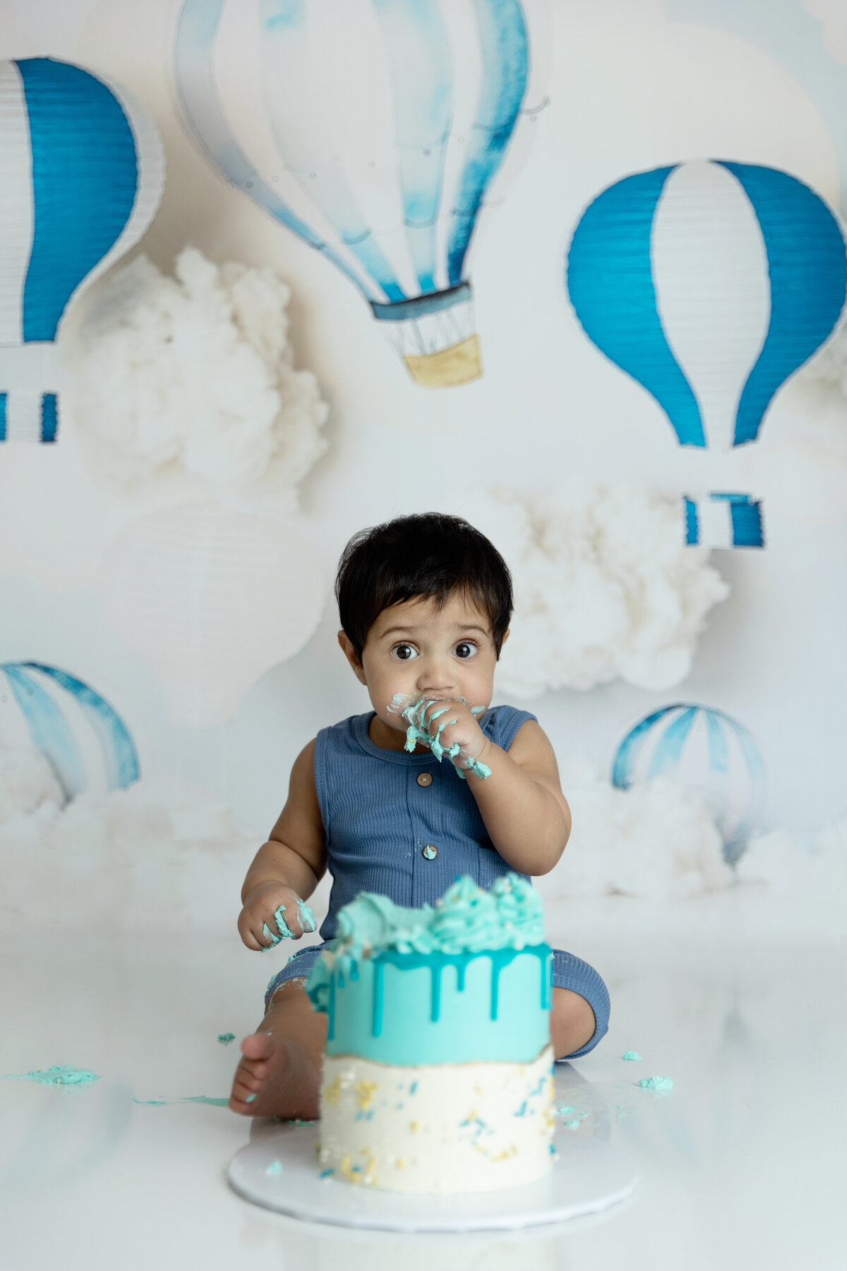 A toddler in a blue onesie makes a silly face while eating cake in a studio