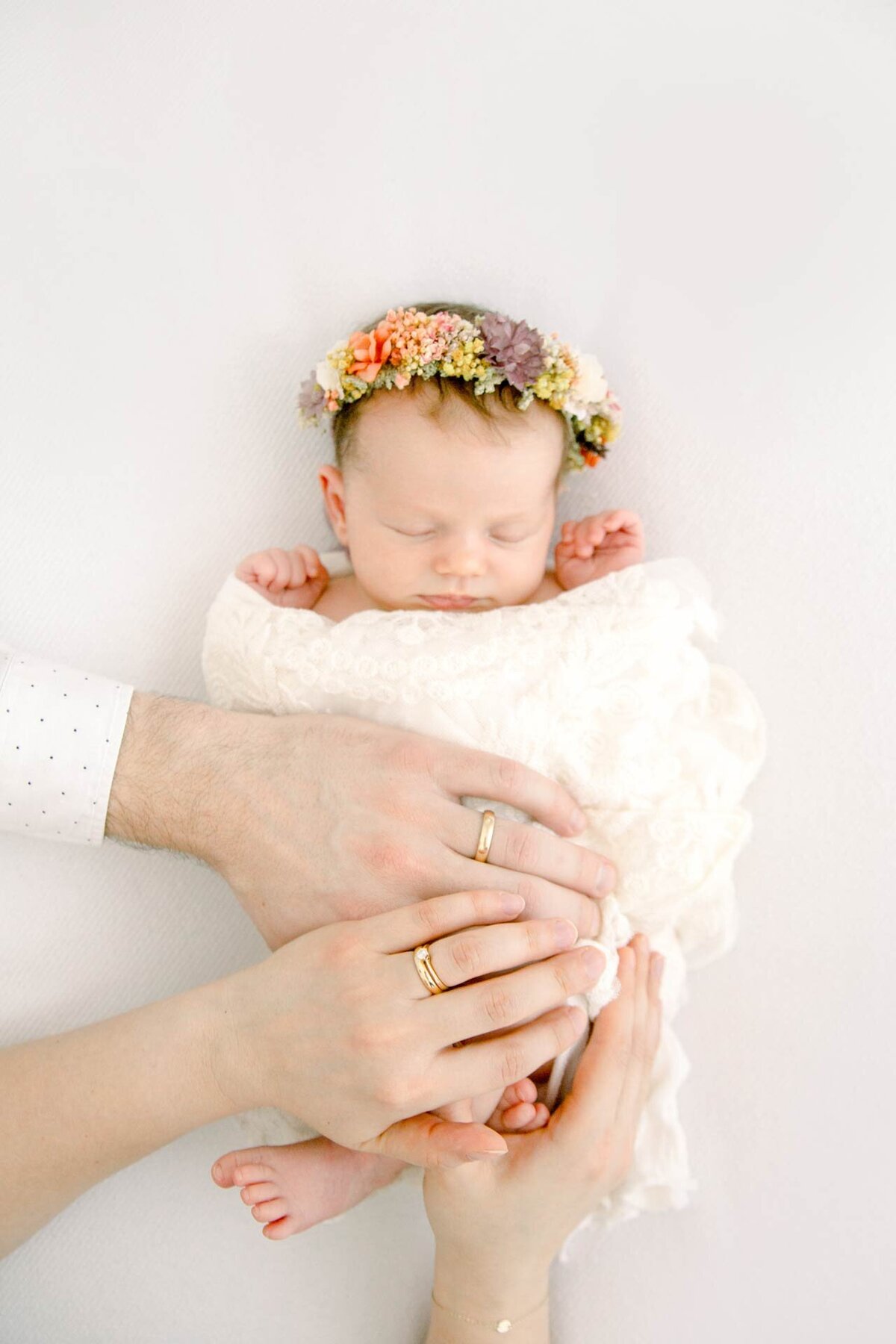 Newborn Fotoshooting mit schlafendem Neugeborenem mit buntem Blumenkranz und auferlegten Händen der Eltern.