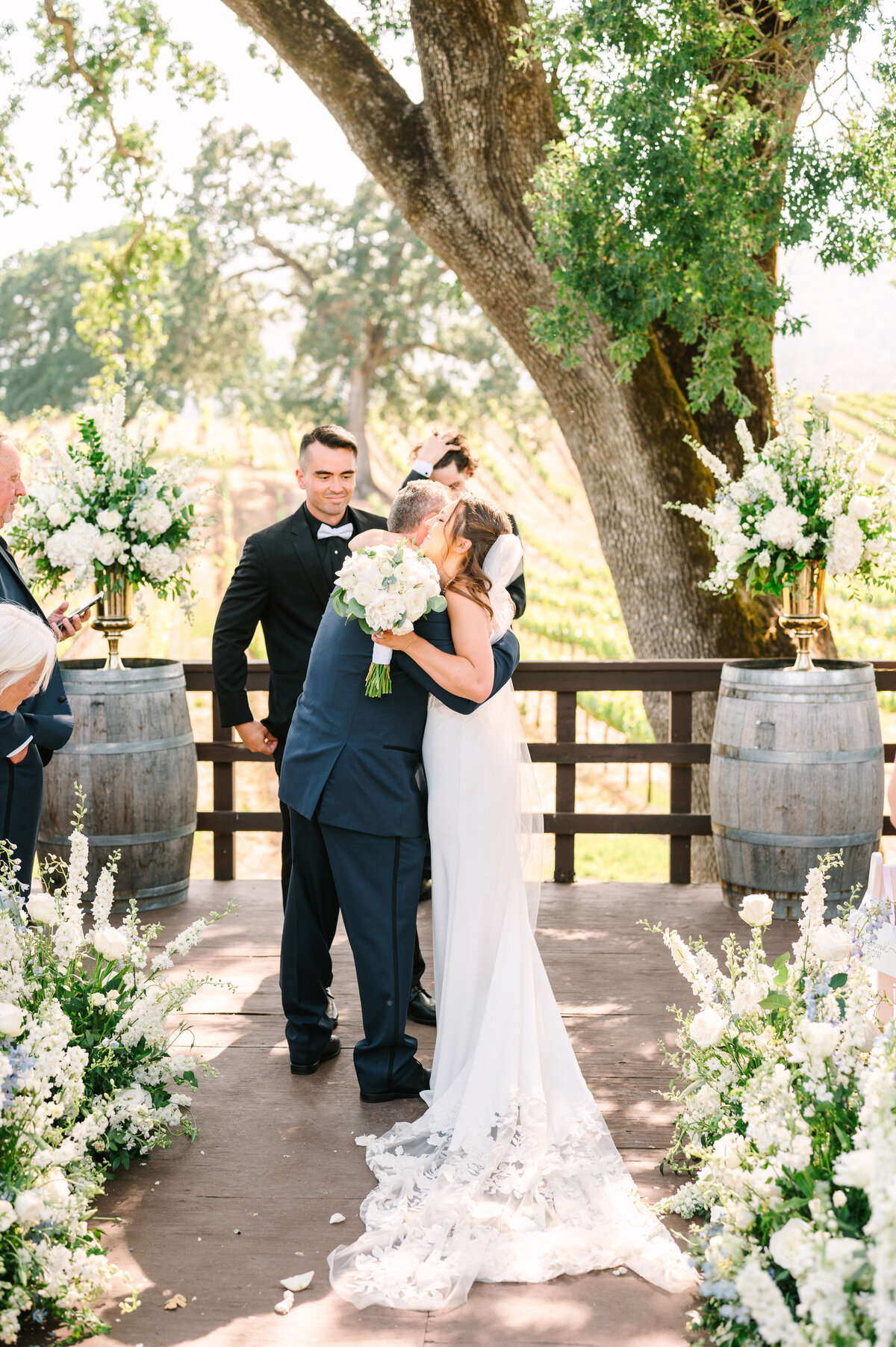 bride hugging her father next to soon-to-be groom in sonoma vineyard before wedding ceremony with white florals and greenery all around.