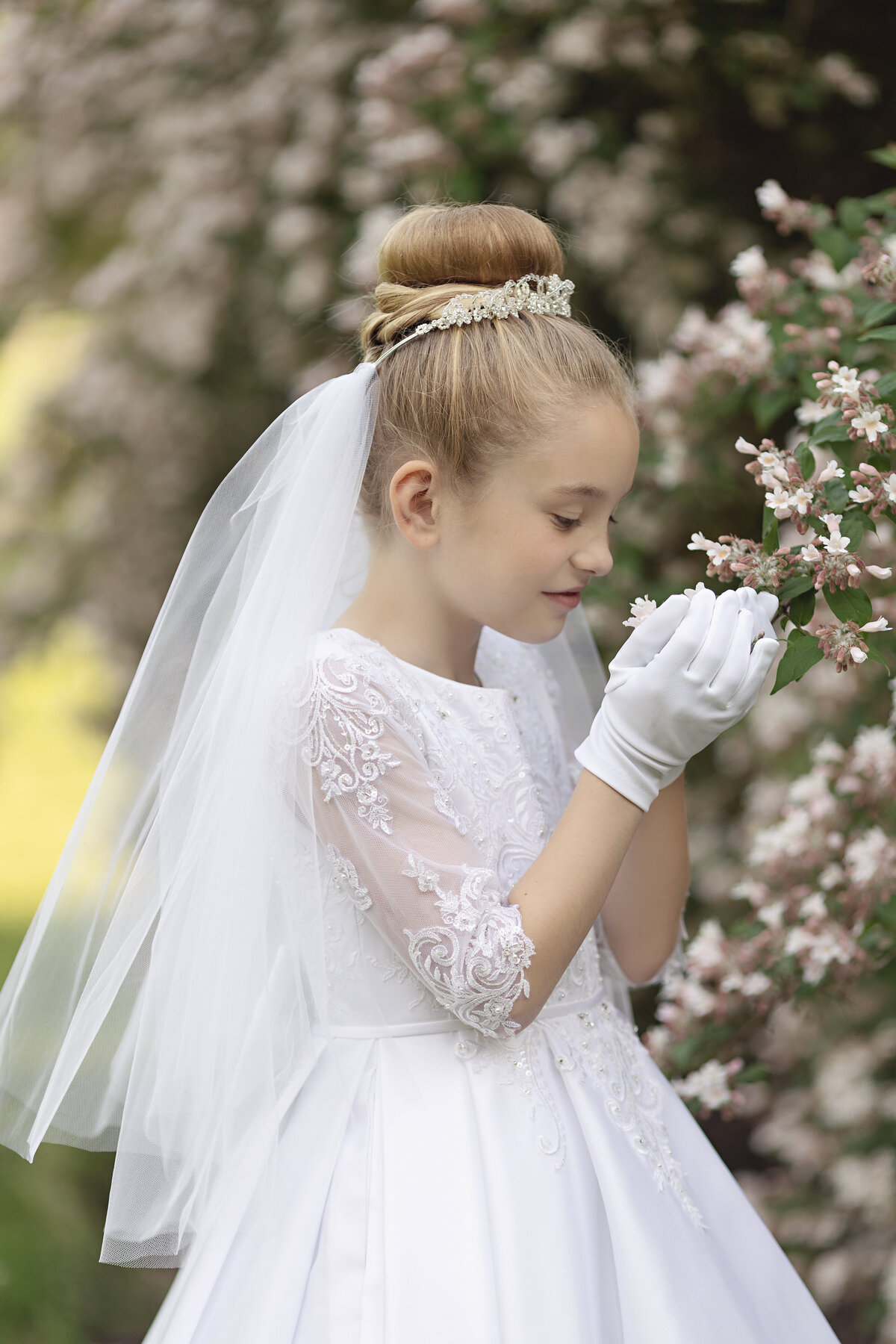 A young girl in a white communion dress and white gloves smells some flowers in a garden