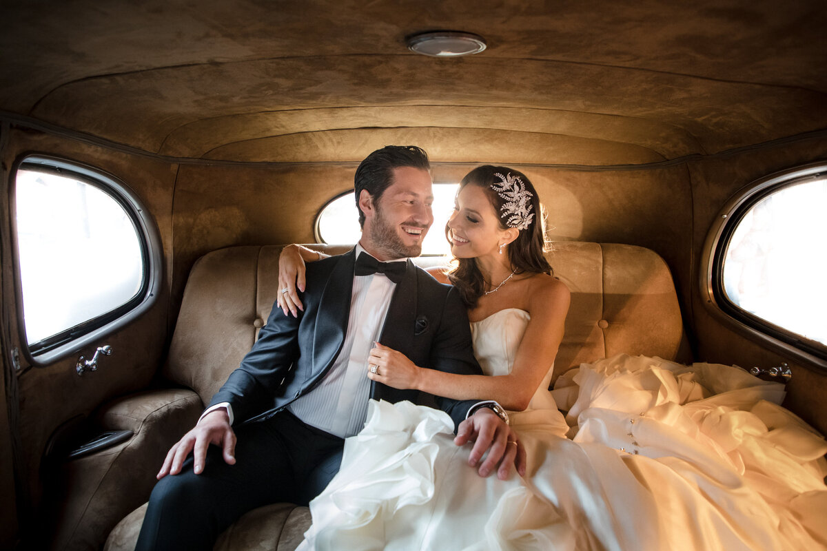 A wedding couple sitting close with their arms around each other in the back of a car.
