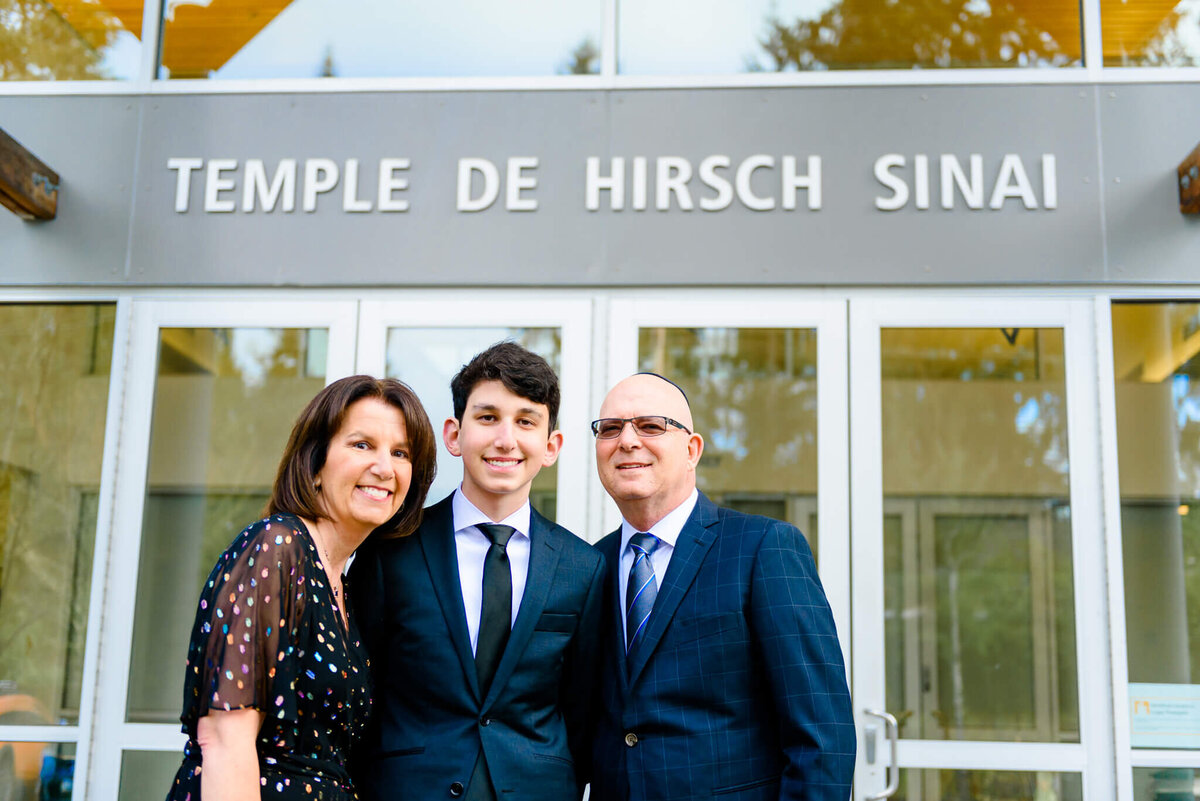 A teenager in a blue suit stands with mom and dad at the temple entrance before his mitzvah