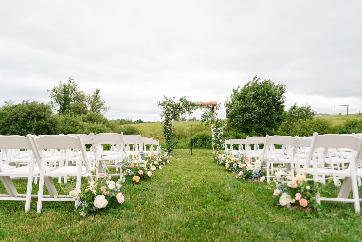 Rows of white chairs facing a floral-decorated archway set up for a wedding ceremony in a grassy field.