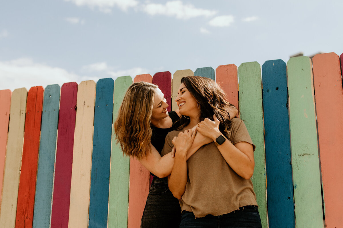 same sex couple hugging in front of a rainbow fence
