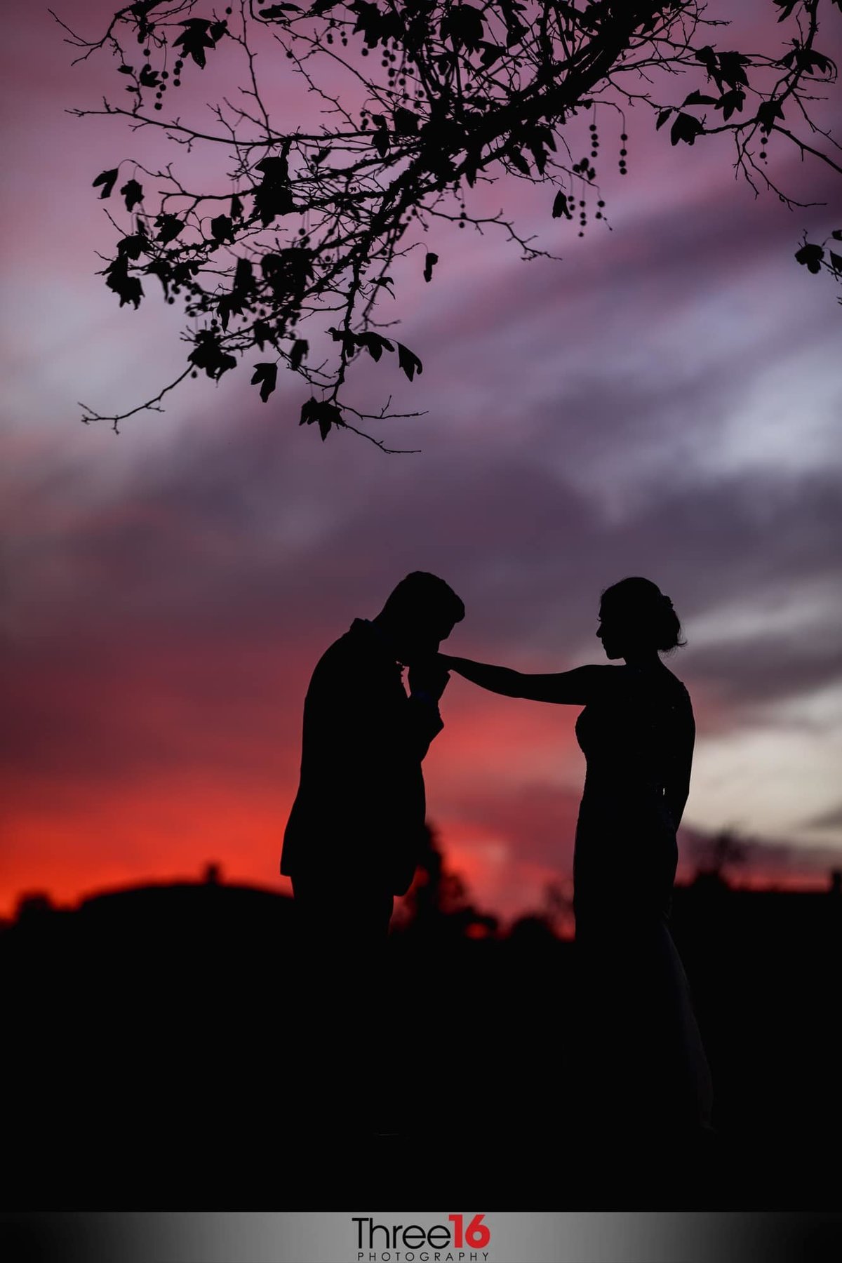 Silhouette photo of Groom kissing his Bride's hand with beautiful red and gray skies behind them