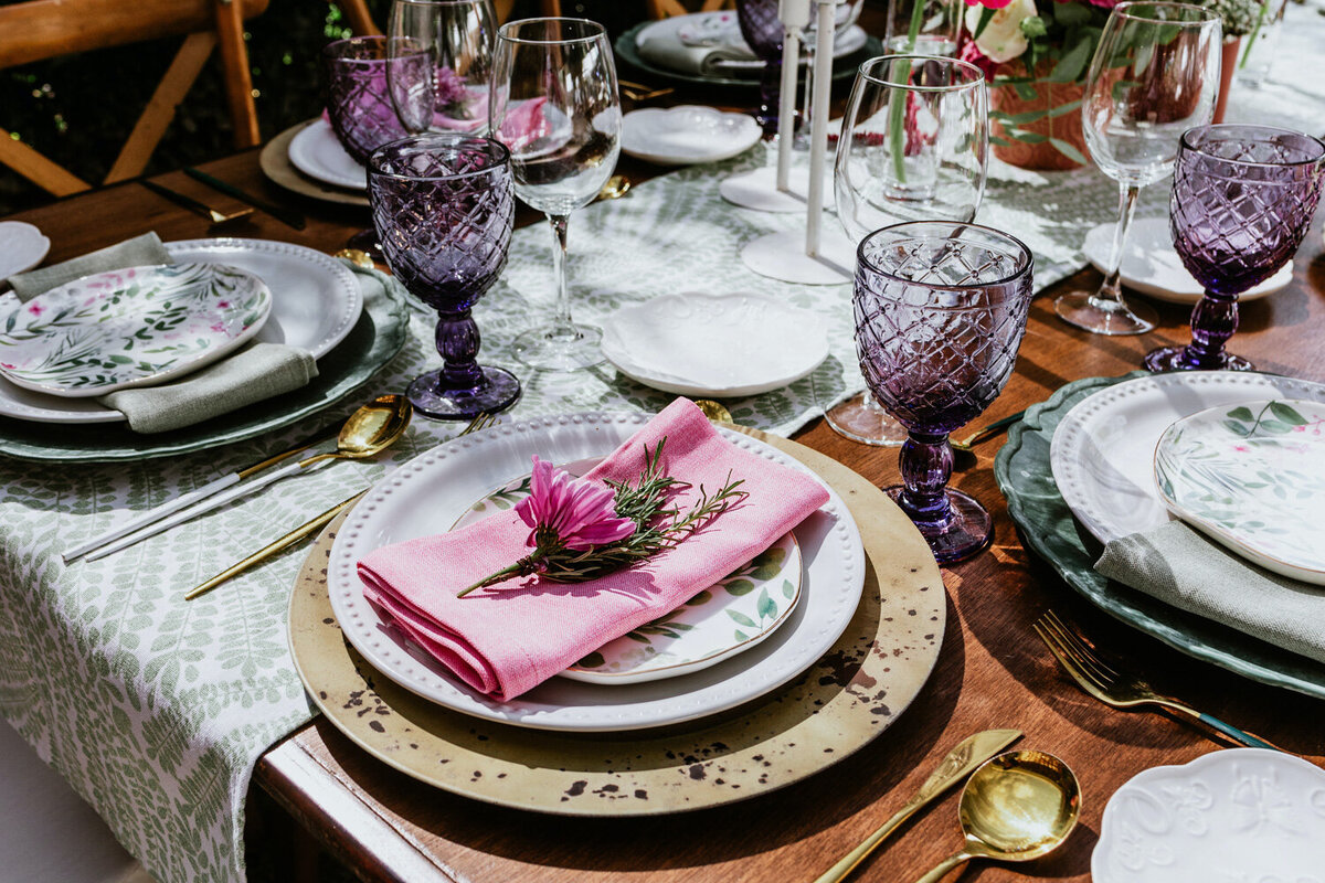 Table with plates and utensils designed with pink table napkin, flowers and purple wine glasses