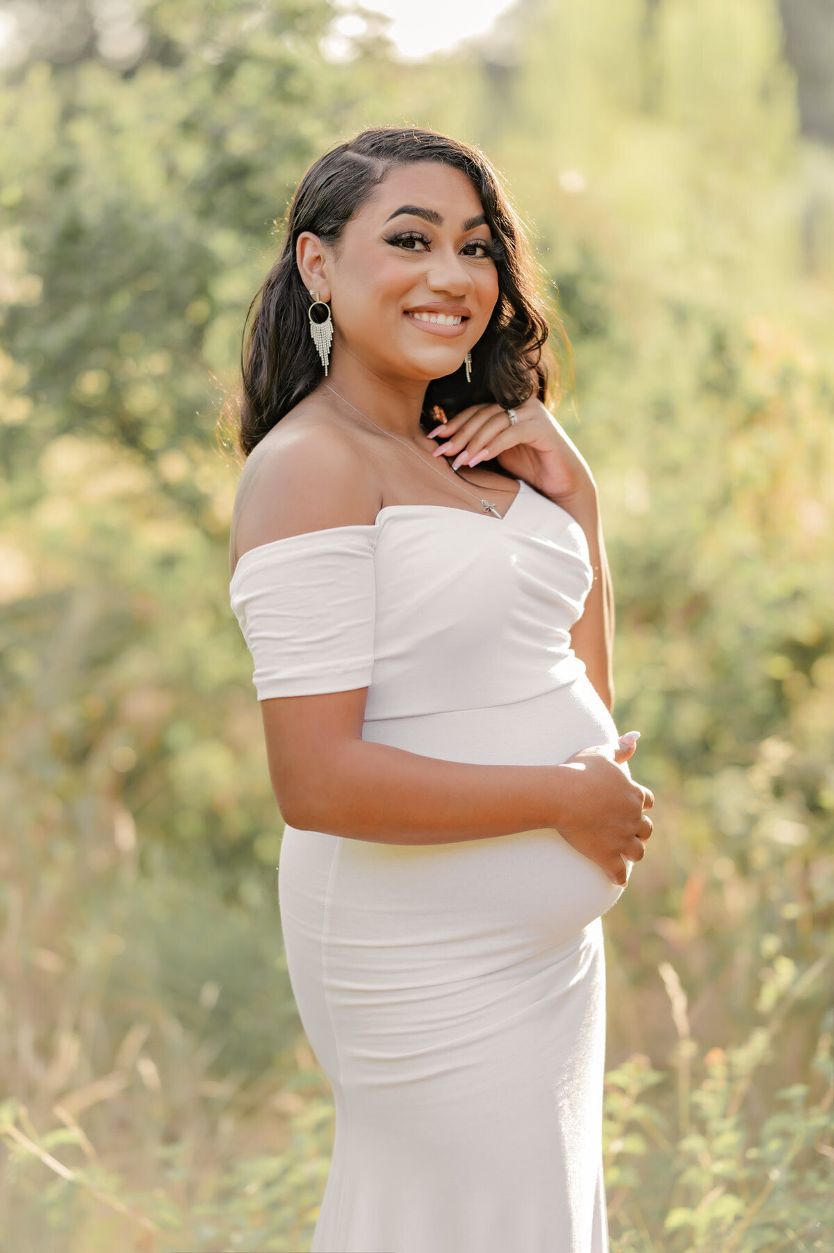 Beauitful glowing portrait of a pregnant woman in a white gown smiling at the camera.