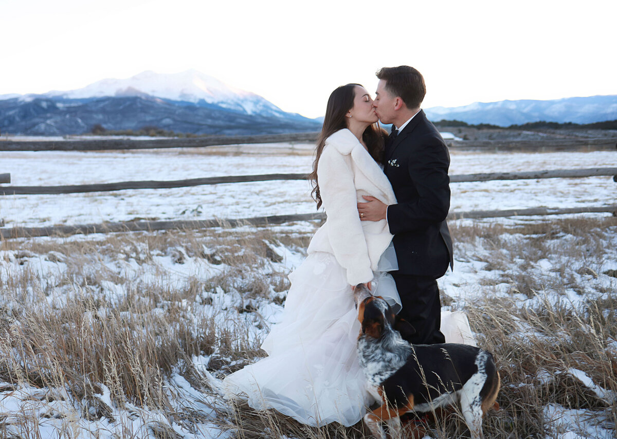 Bride and groom kissing each other, with a snowy mountain view and a beautiful sunset.