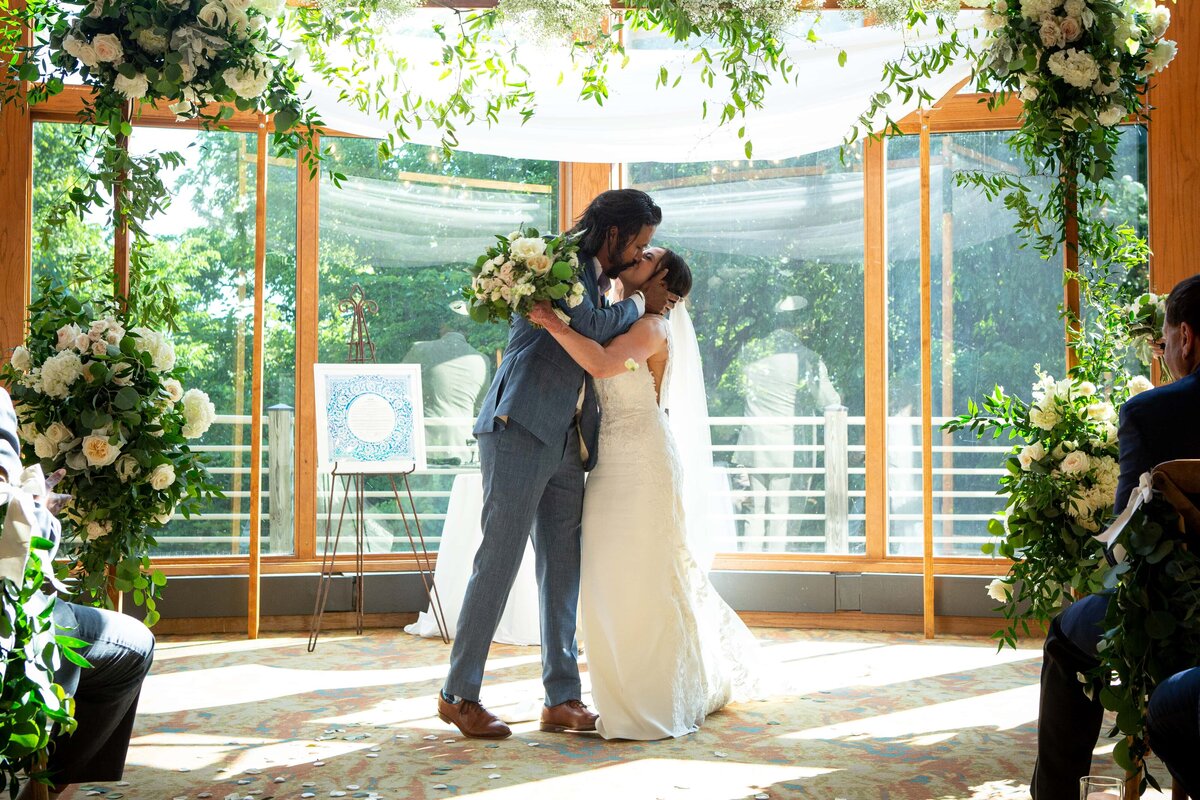 A bride and groom kiss passionately at the altar under a floral arch, in a sunny room surrounded by seated guests at an Iowa wedding.