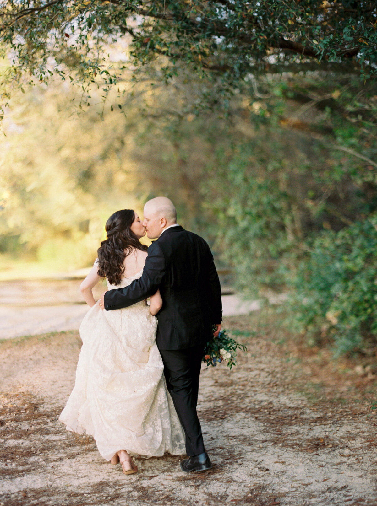 Bride and groom kiss as they walk down a tree lined path