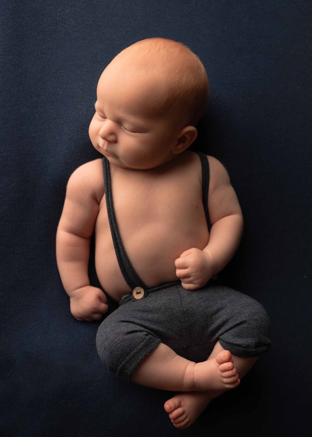Maddie Rae Photography baby sleeping on a navy blue background. he is wearing little blue suspender pants. his feet are crossed