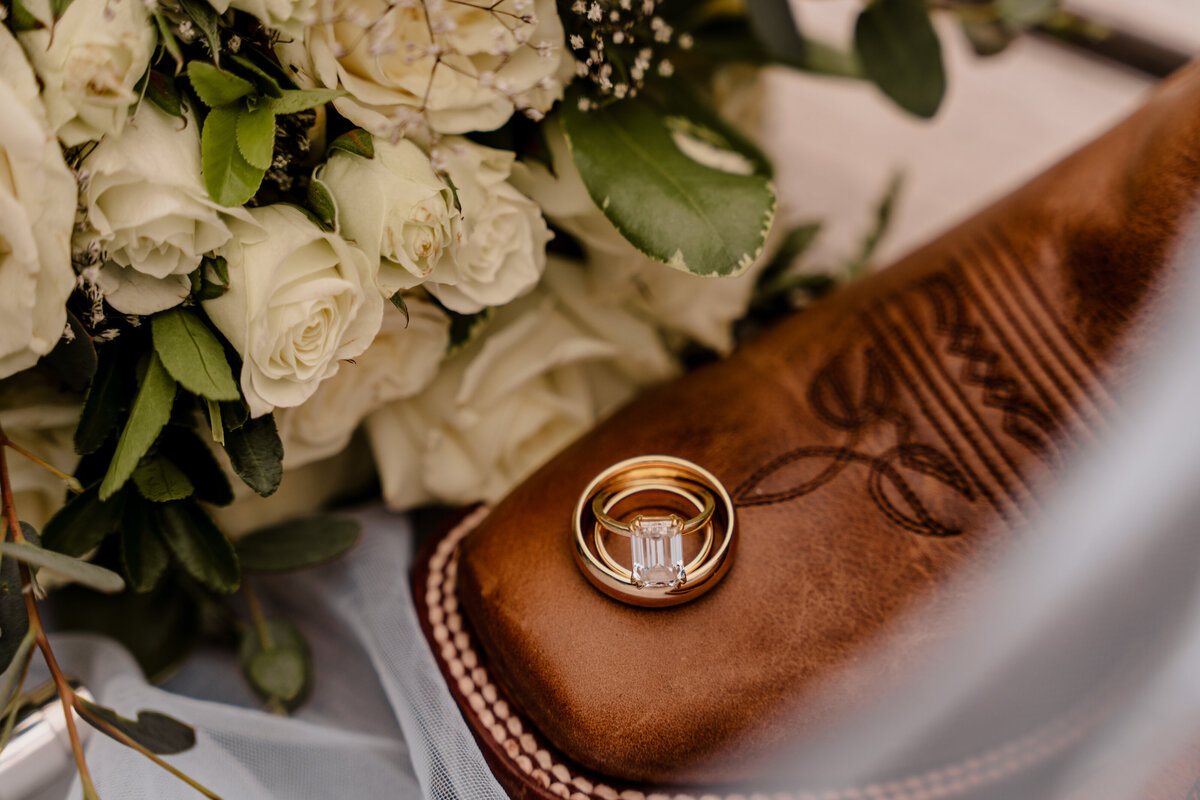 An emerald cut wedding ring is displayed sitting atop a cowboy boot and wedding bouquet.