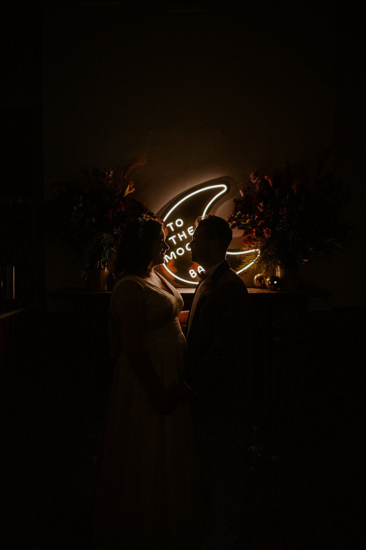 A couple are silhouetted by their neon sign that reads "to the moon and back". They ceremony was at The Asylum Chapel in London and they and their guest boarded a vintage style red London bus to a resturant in Camberwell called The Camberwell Arms.