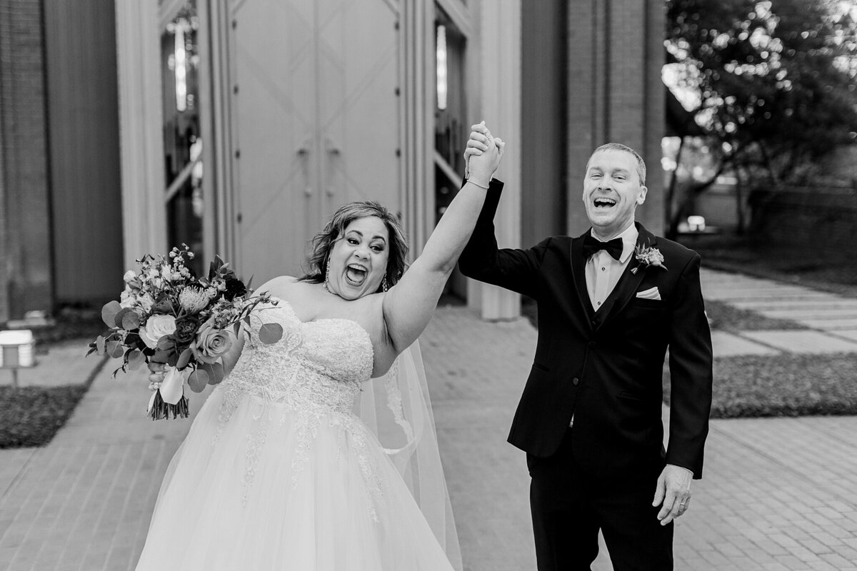 A black and white portrait of a bride and groom joyfully celebrating after their wedding ceremony at the Marty Leonard Community Chapel in Fort Worth, Texas. They bride is on the left and is wearing a detailed, intricate, short sleeve, white dress with a long veil and is holding a bouquet. The groom is on the right and is wearing a black tuxedo with a bowtie and boutonniere.