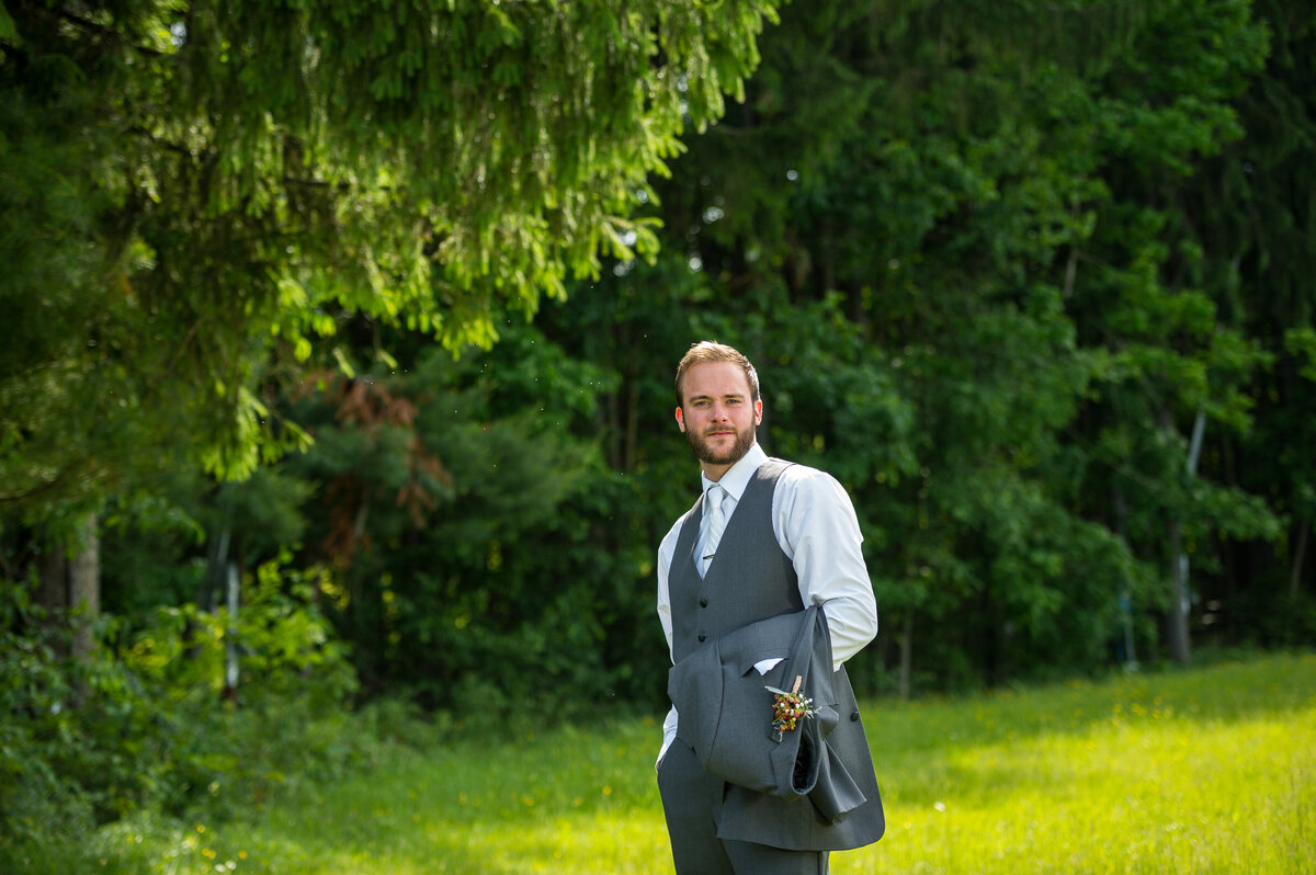 Groom holding jacket standing in the woods.